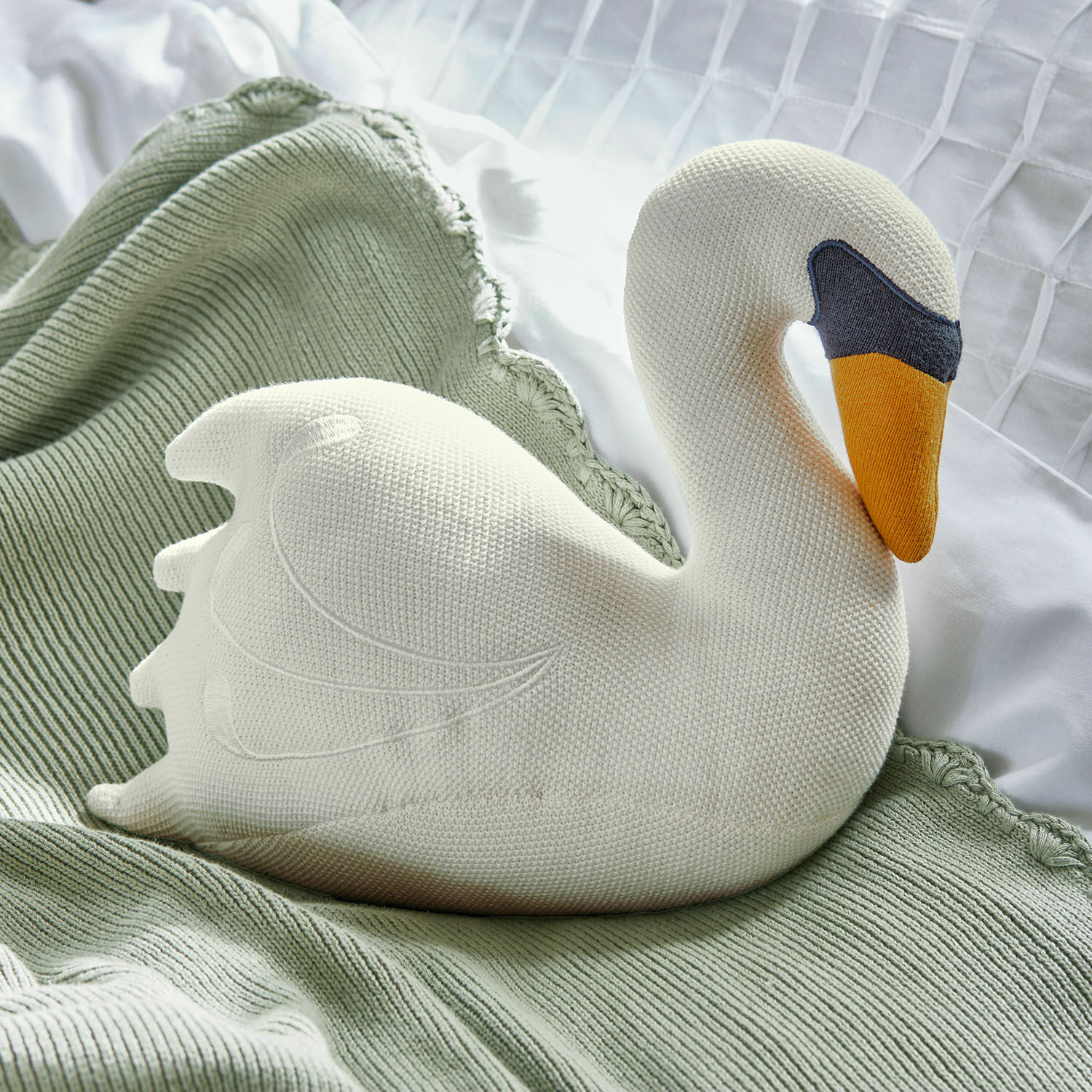 Peluche Cariella Coussin cygne blanc en tricot posé sur un plaid vert, décoration douce pour chambre