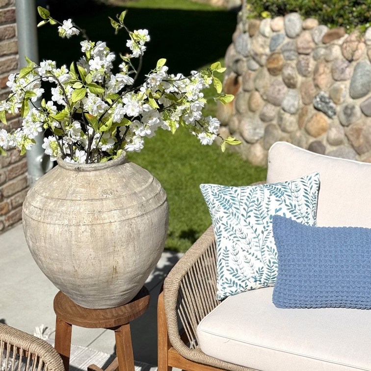 Grand vase en céramique beige avec fleurs blanches sur un support en bois à côté d'un canapé avec coussins dans un jardin