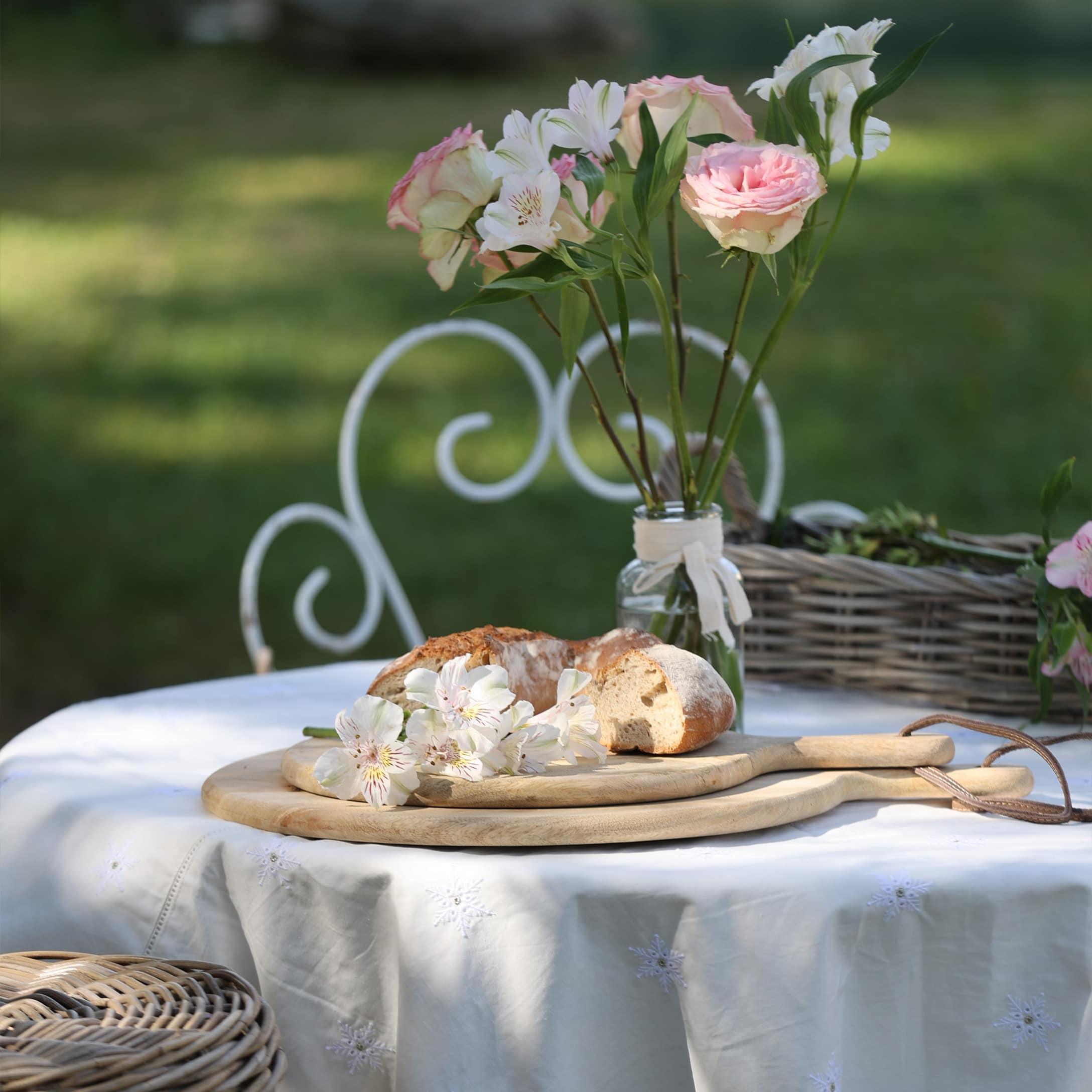 Planche à découper en bois sur nappe blanche avec pain et fleurs dans un vase en verre au jardin