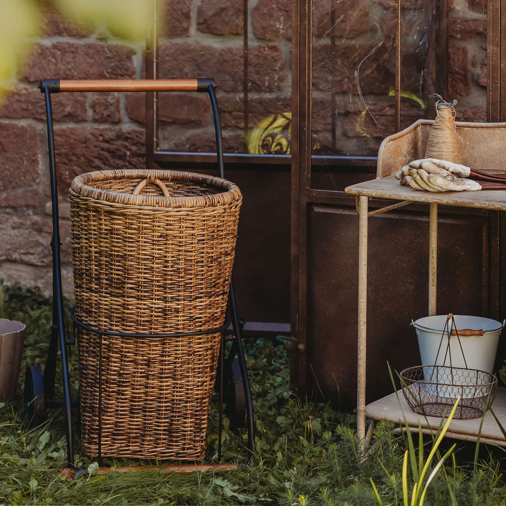 Chariot avec panier en osier et cadre métallique sur l'herbe près d'un mur en briques, outils de jardin à côté