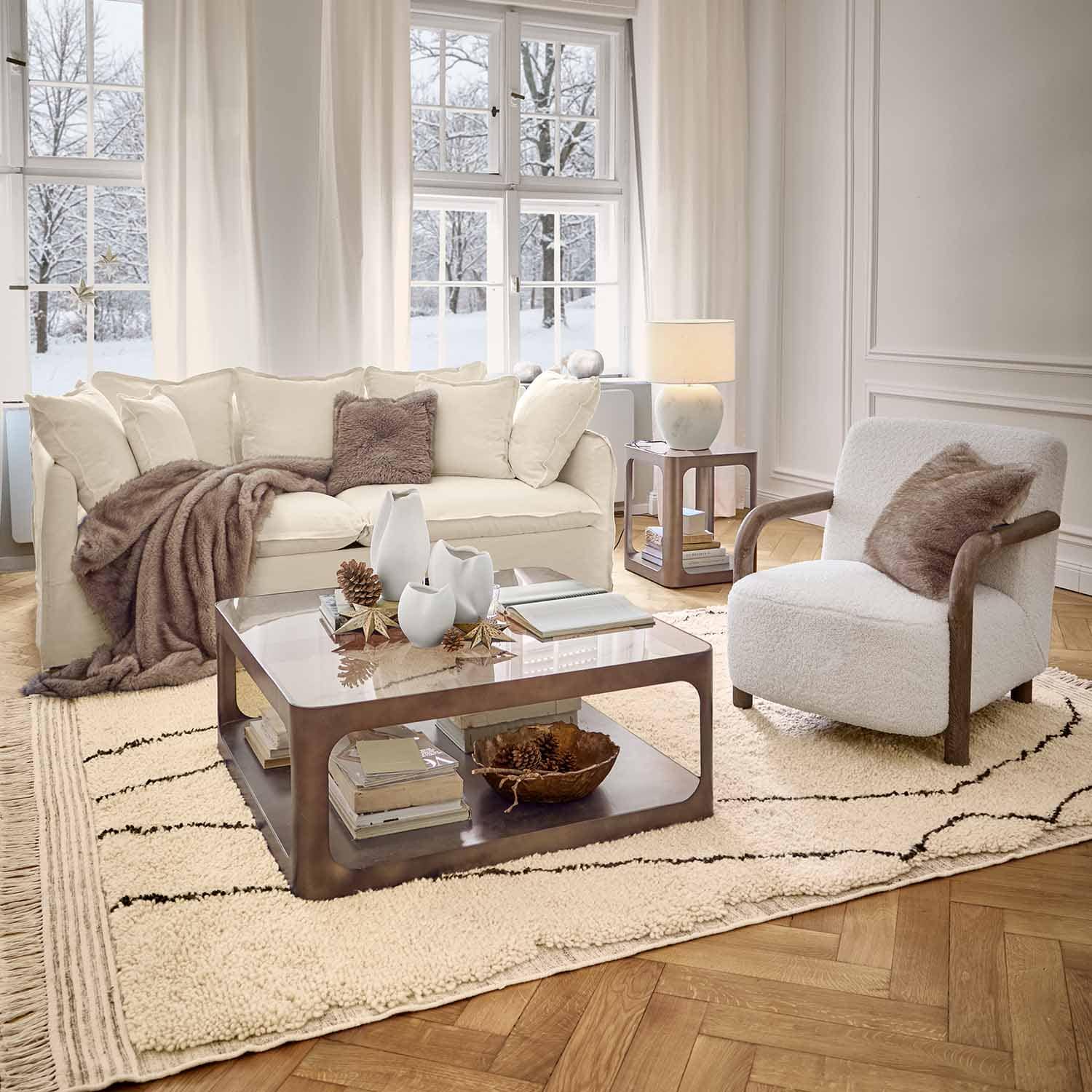 Salon chaleureux avec canapé blanc, fauteuil en bois et tapis moelleux dans une ambiance hivernale