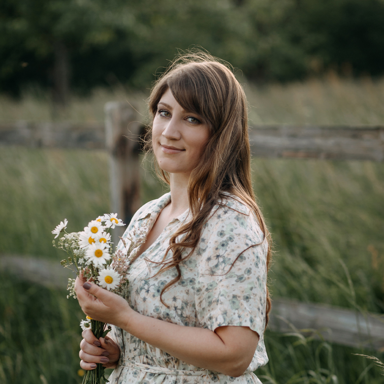 Femme en robe fleurie tenant un bouquet de marguerites devant un champ vert et une clôture en bois