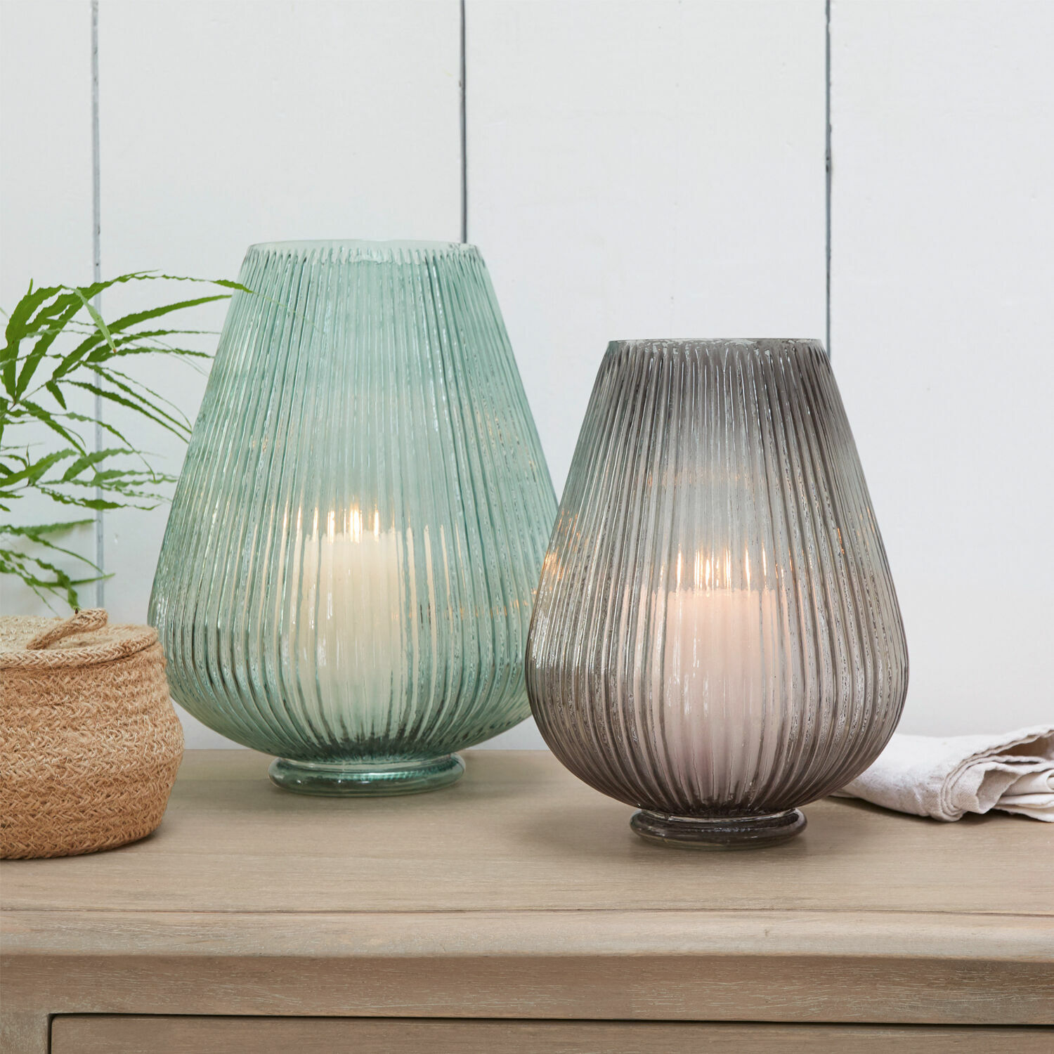 Lot de 2 vases Fenis Deux lanternes en verre vert et gris avec bougies blanches sur une table en bois clair