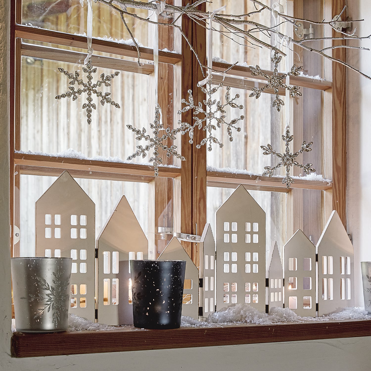 Décoration hivernale avec maisons en bois blanches, photophores et flocons suspendus sur le rebord de fenêtre en bois