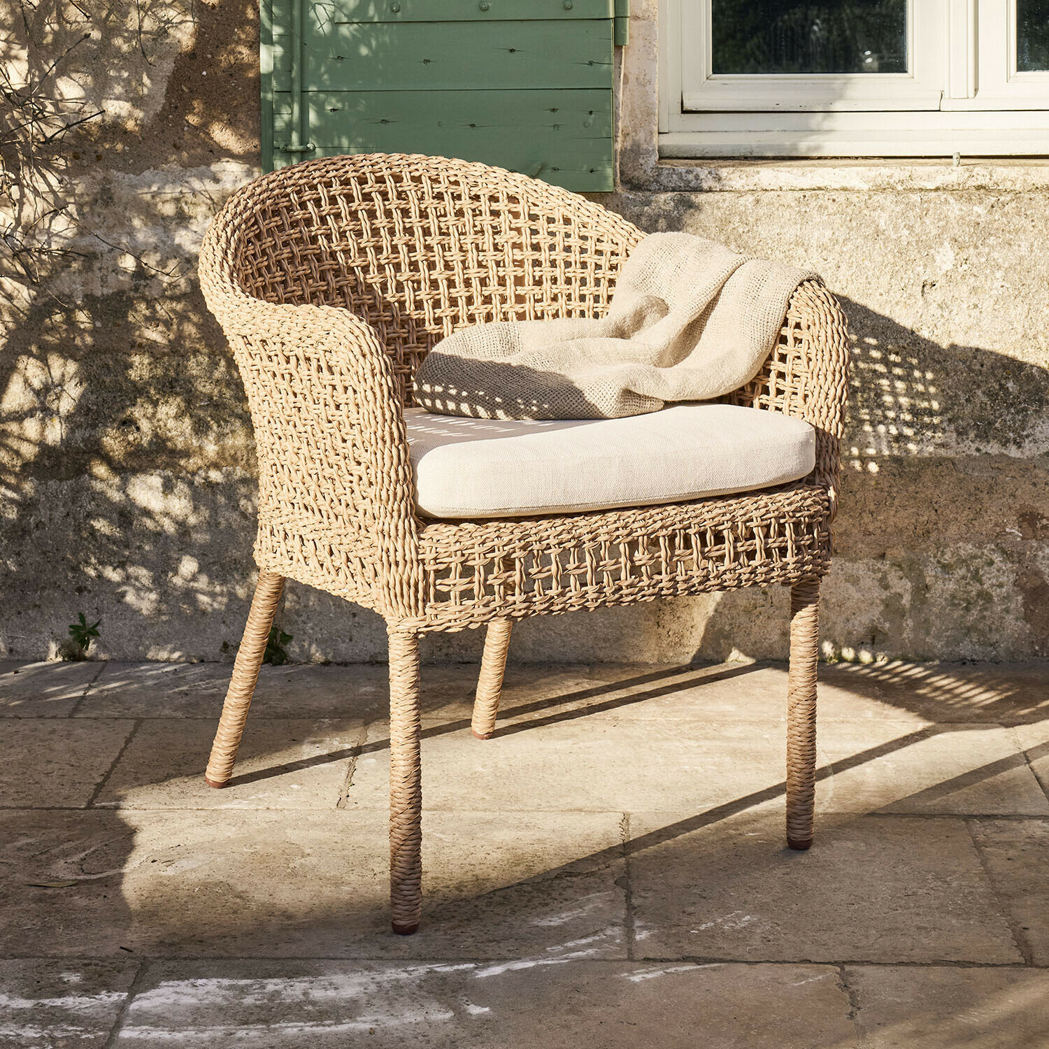 Fauteuil en rotin tressé naturel avec coussin clair et plaid beige sur terrasse en pierre