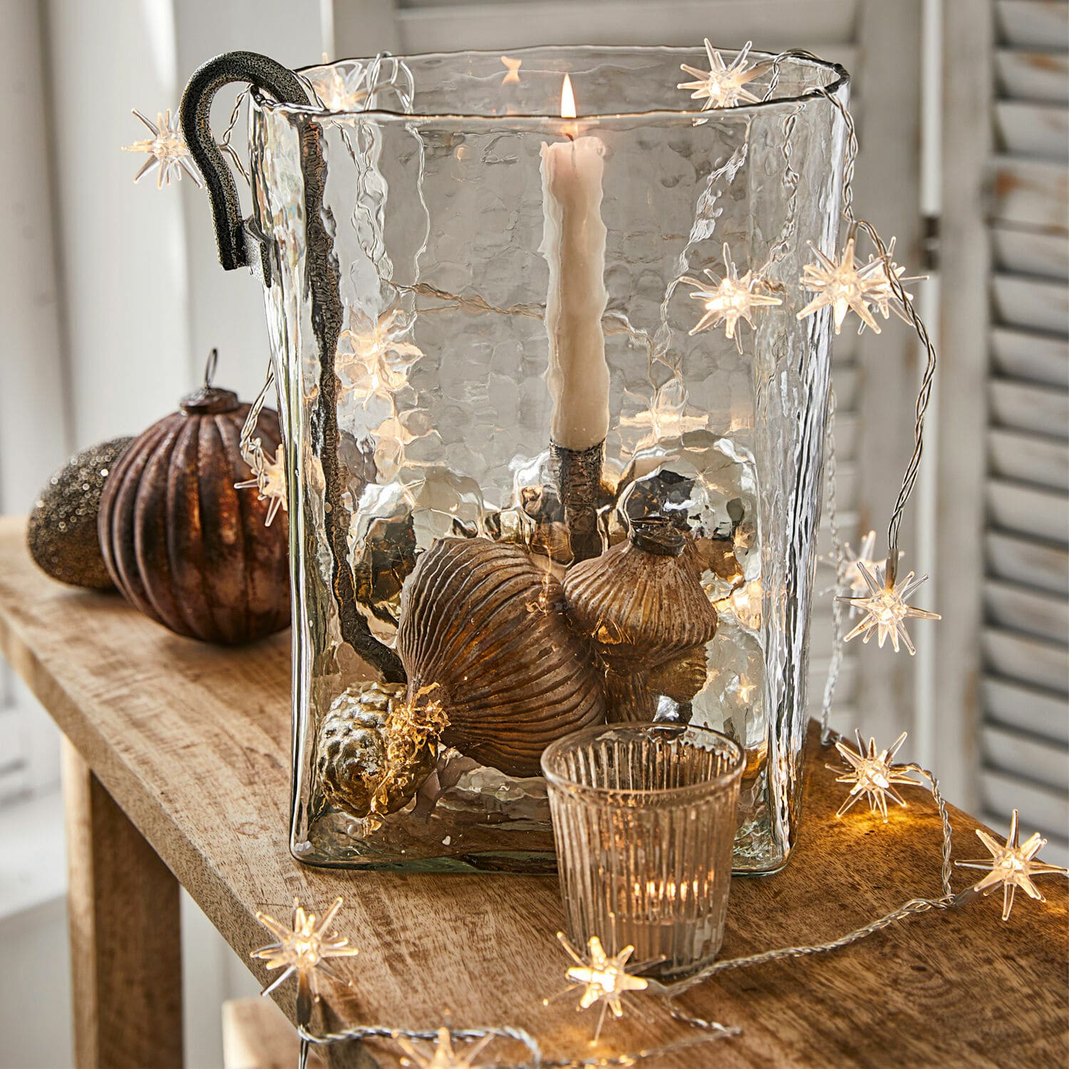 Guirlande lumineuse Nelisse Bougeoir en verre avec poignée en métal et décorations cuivre sur table en bois avec guirlande lumineuse étoile