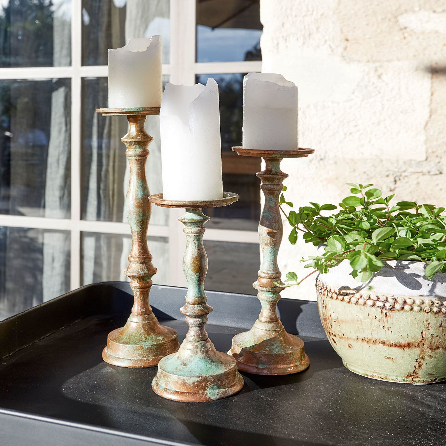 Lot de 3 bougeoirs Floirac Trois bougeoirs en laiton avec bougies blanches partiellement fondues sur un plateau noir à côté d'une plante verte