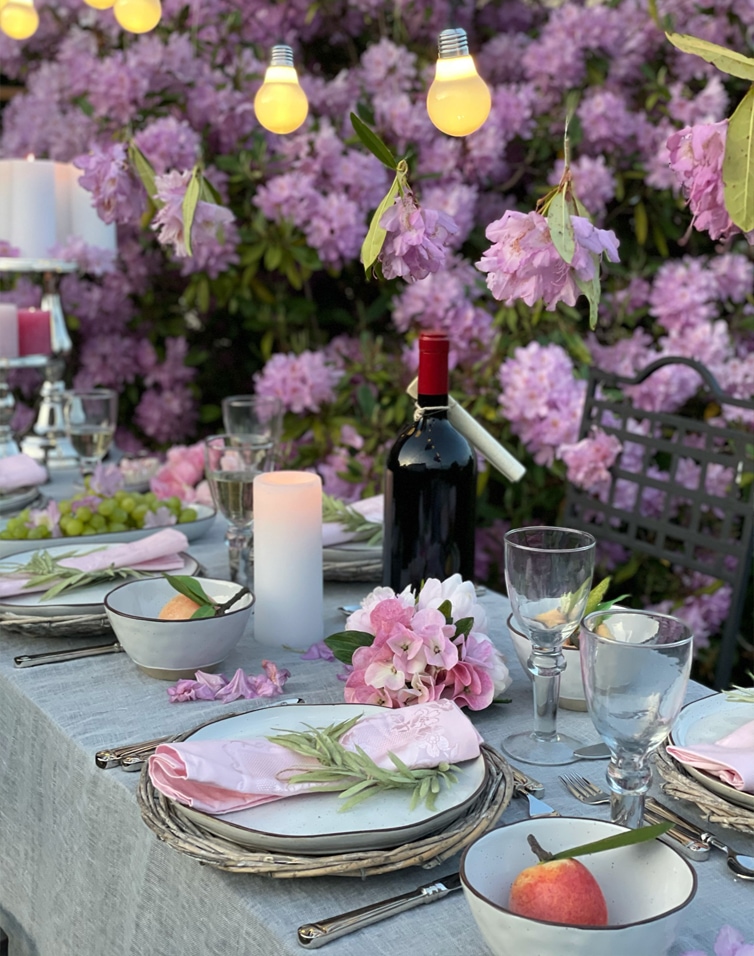 Table d'été décorée avec fleurs roses, guirlandes lumineuses et bouteille de vin devant des buissons violets.