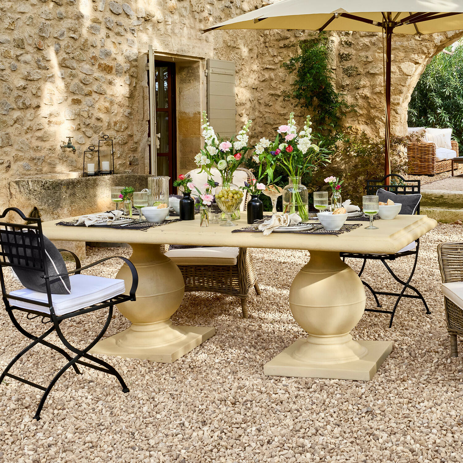 Table Talvero Grande table de jardin beige en pierre sur patio gravillonné avec fleurs et chaises en métal et osier