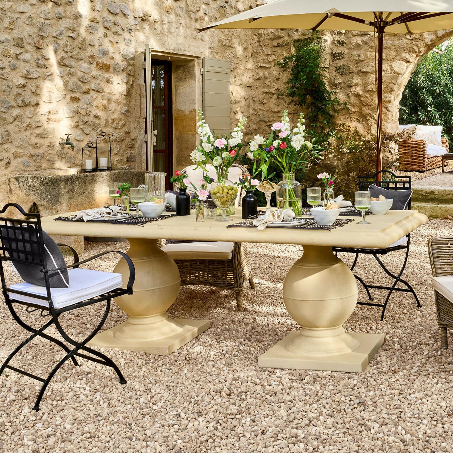 Table Talvero Grande table de jardin beige en pierre sur patio gravillonné avec fleurs et chaises en métal et osier