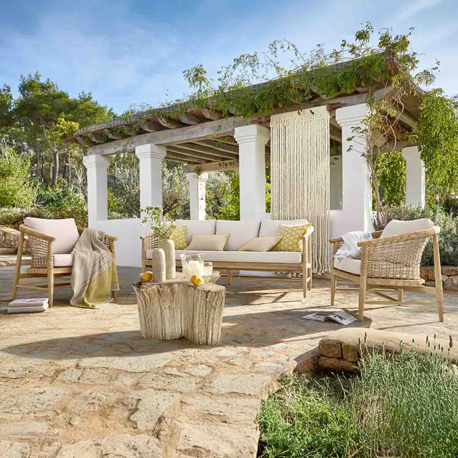Ensemble de mobilier de jardin en bois clair avec coussins crème sur terrasse en pierre sous une pergola végétalisée