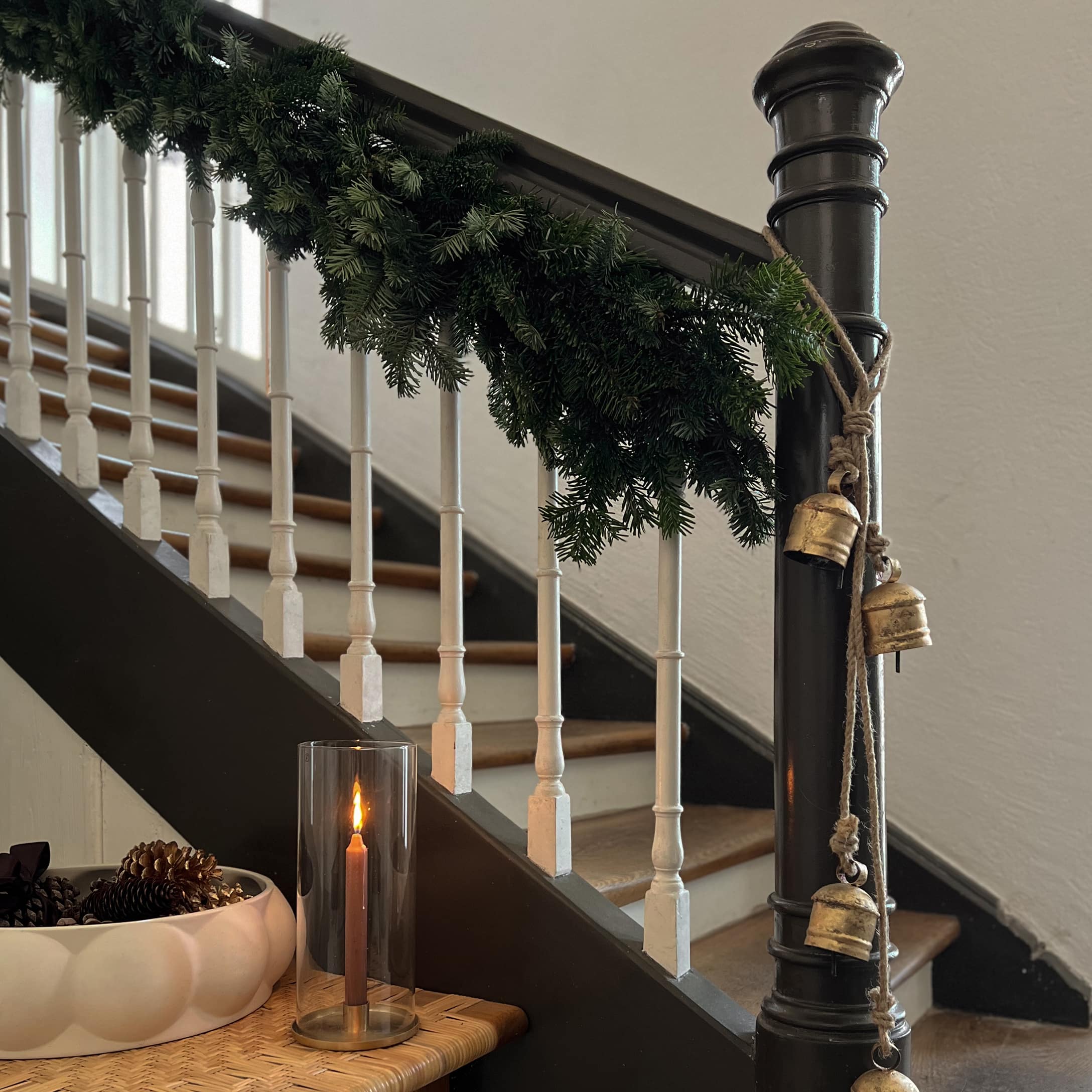 Décoration hivernale avec guirlande verte, cloches dorées et bougie dans un photophore en verre près de l'escalier