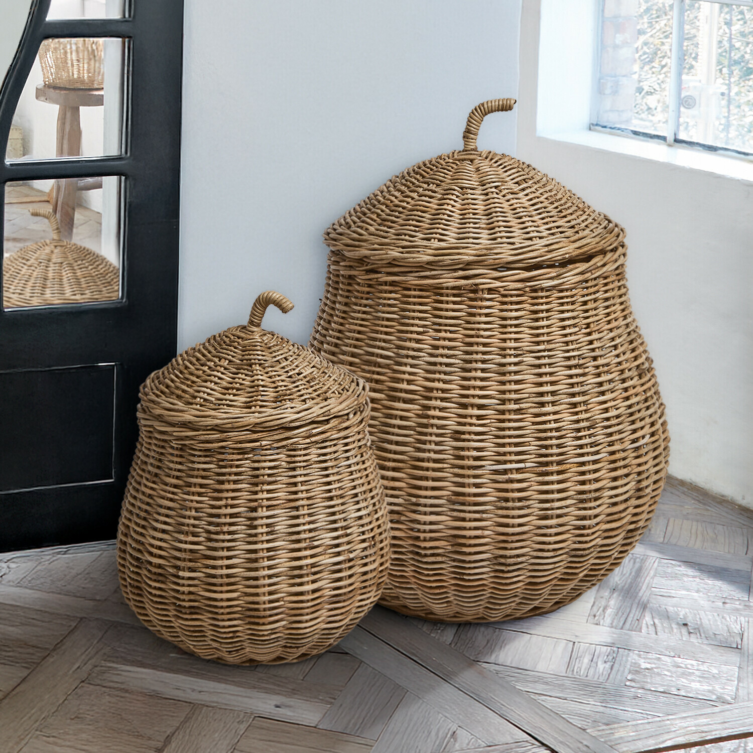 Lot de 2 paniers Fernn Deux paniers en osier naturel avec couvercles posés près d'une fenêtre sur un sol en parquet