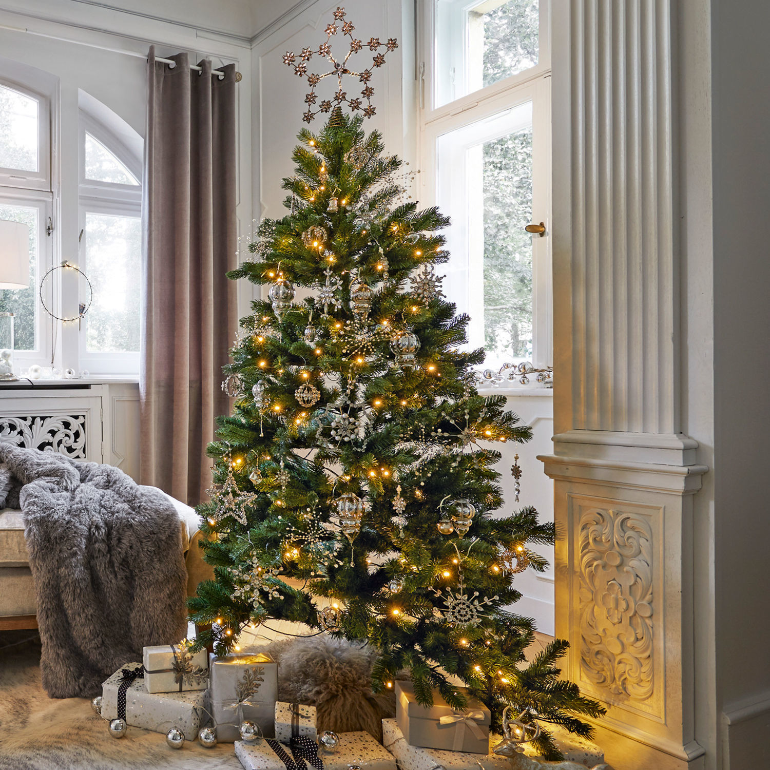Lot de décorations de Noël Bjouterie Sapin de Noël vert décoré d'ornements argentés et de guirlandes lumineuses dans un salon clair