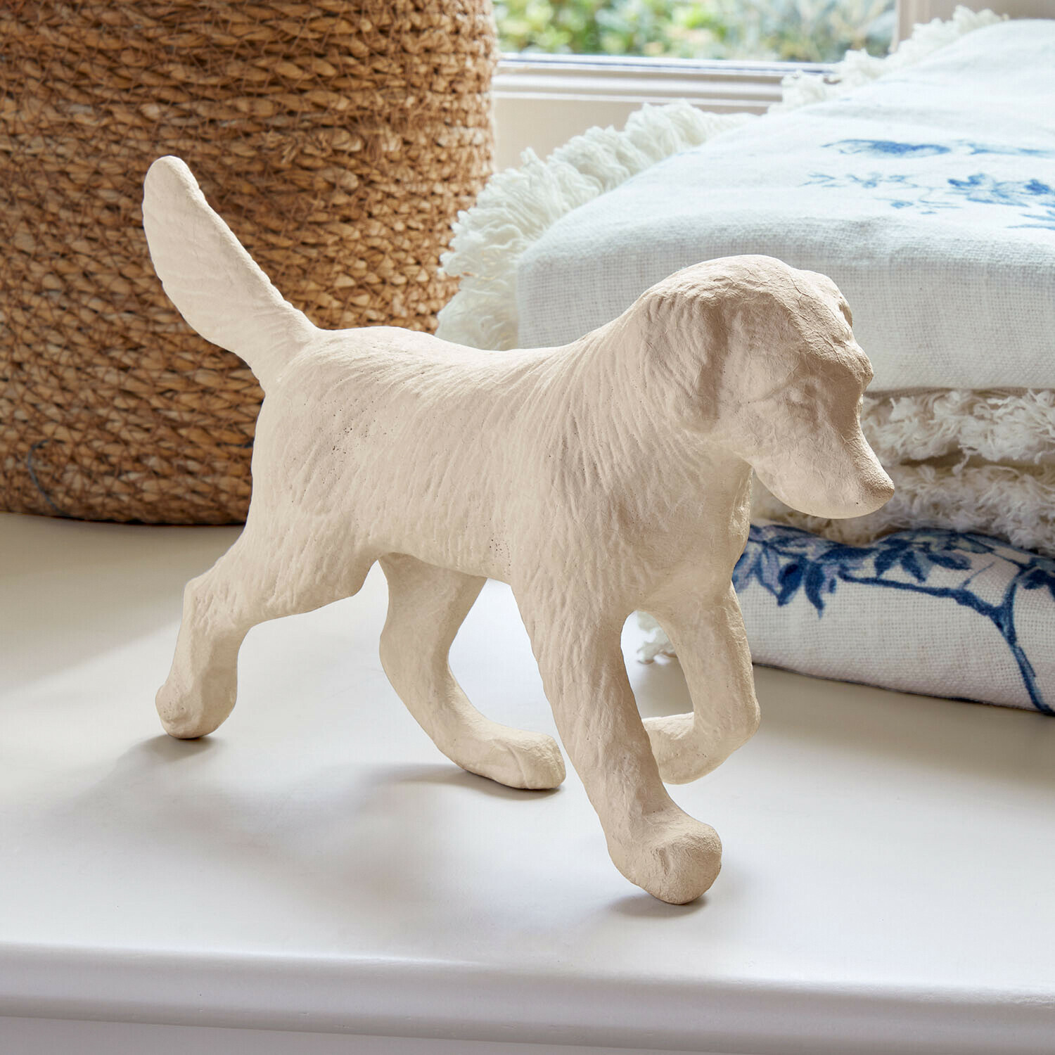 Figurine de chien beige en matériau texturé posée sur une surface blanche avec tissu décoratif en arrière-plan
