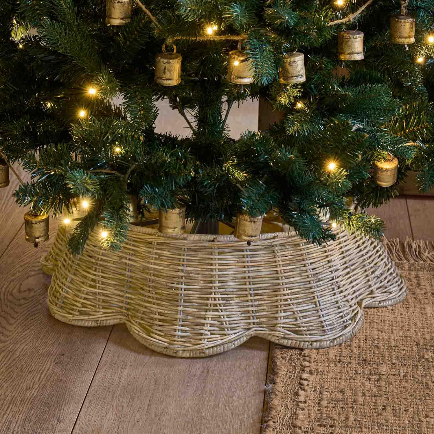Support de sapin en osier naturel beige clair sous un sapin vert décoré de petites lumières chaudes