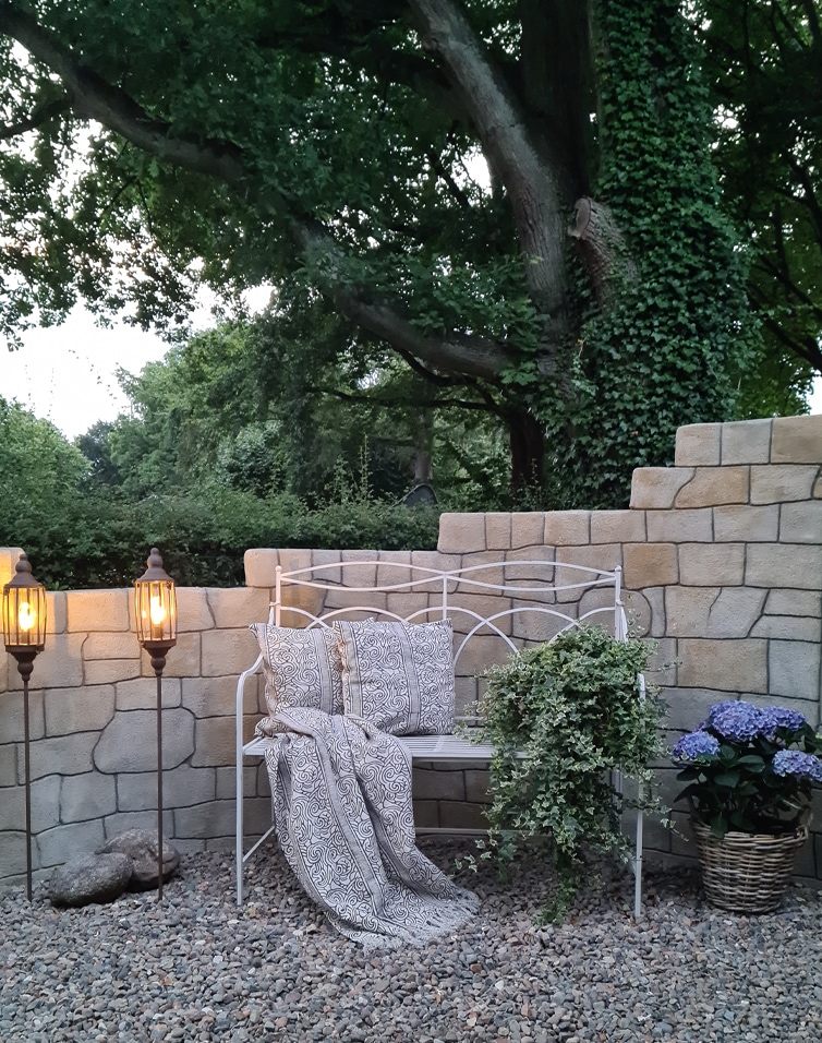 Banc de jardin en métal blanc avec coussins et couverture, entouré d’un mur en pierre et de plantes