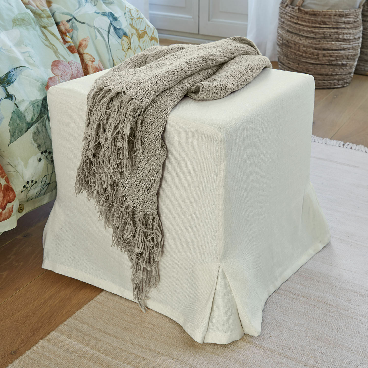 Pouf Ulysette Pouf beige en tissu avec couverture tricotée grise posé sur un sol en bois dans un intérieur chaleureux