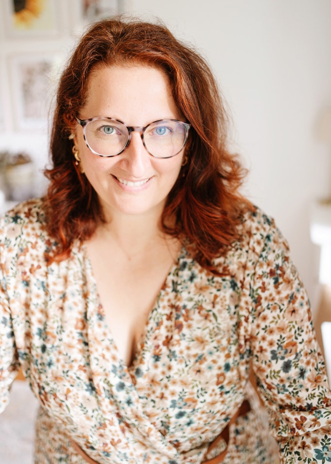 Portrait d'une femme souriante aux cheveux roux et lunettes portant un chemisier fleuri aux tons clairs