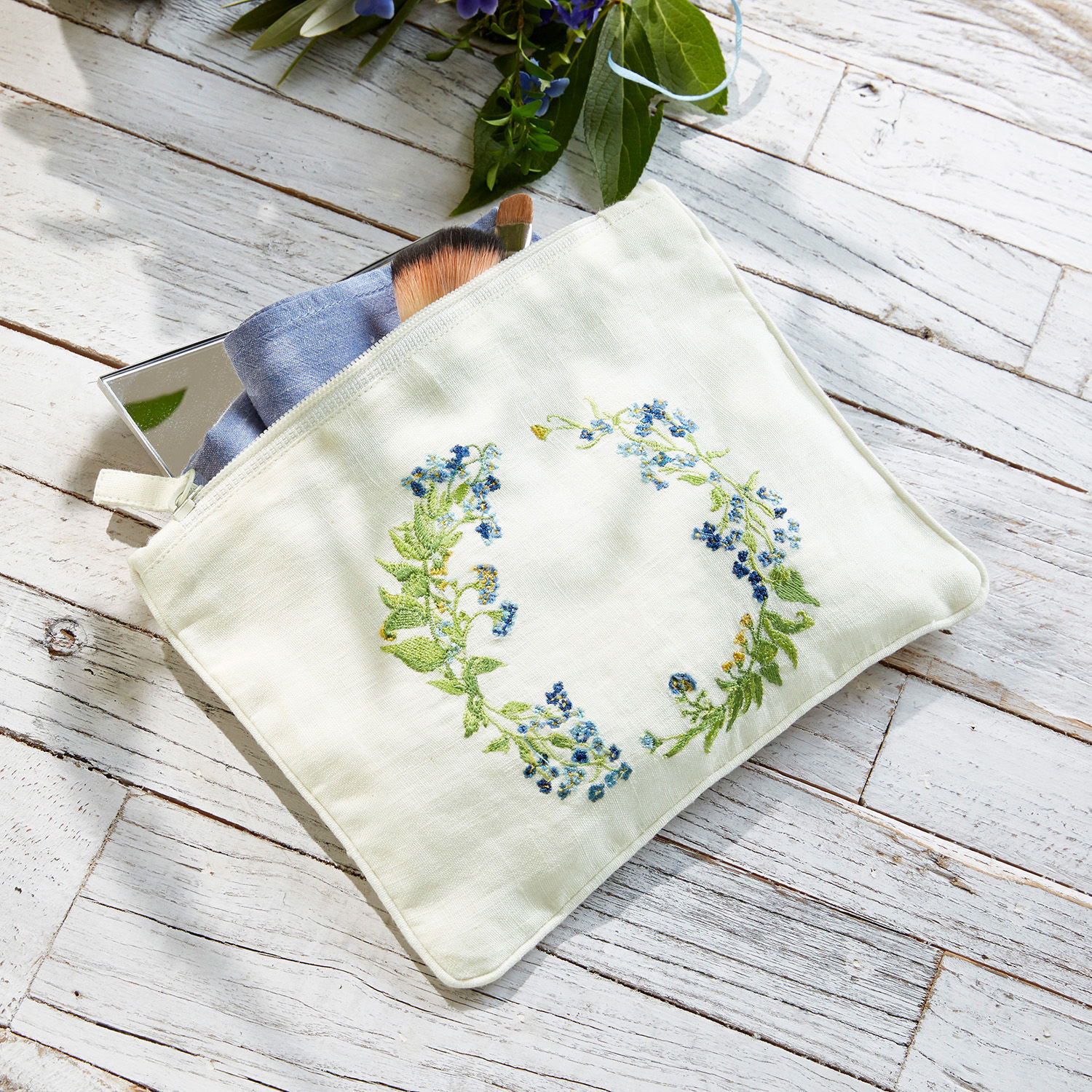 Pochette Fierolla Trousse de maquillage en lin blanc avec broderie florale bleue posée sur un fond en bois clair