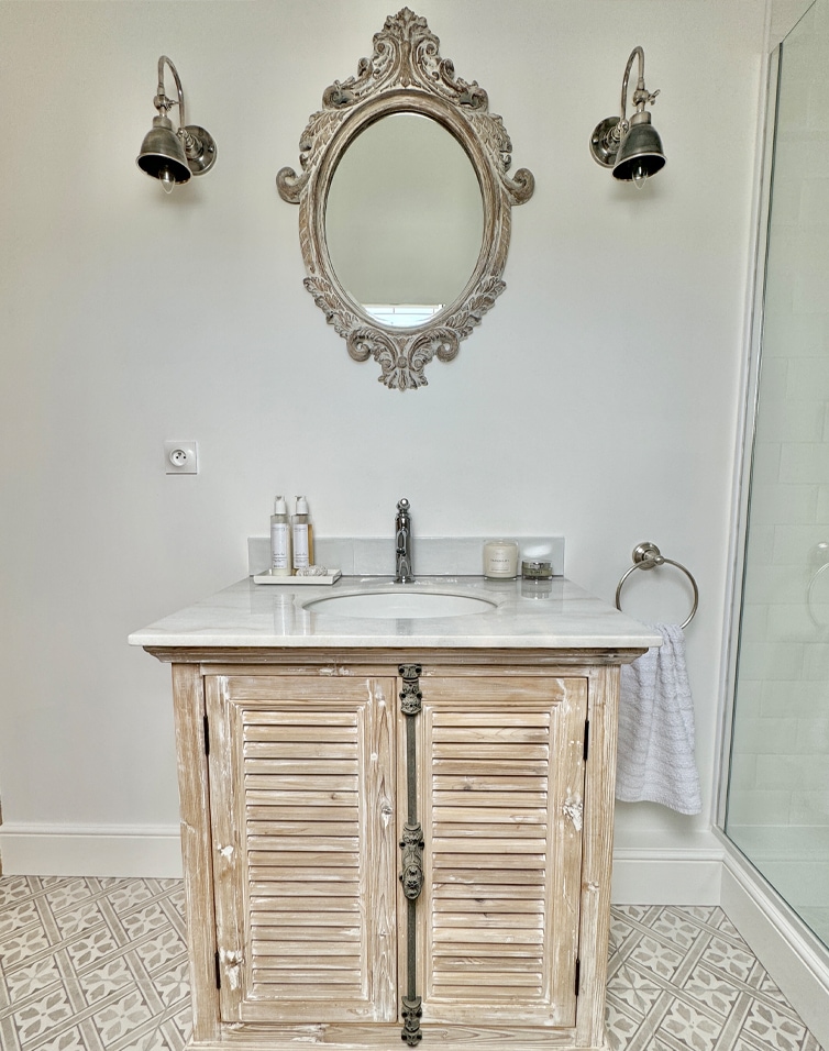 Salle de bain avec meuble en bois patiné, plan en marbre blanc et miroir ovale décoratif vintage