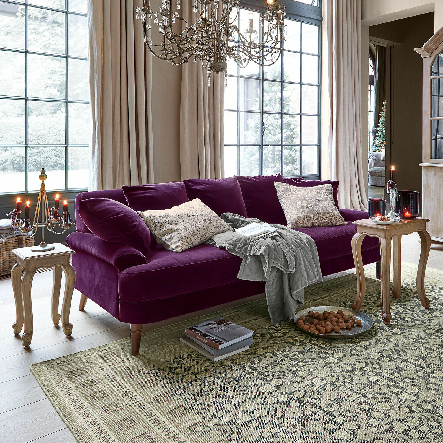 Canapé Rivermere Canapé violet en velours avec coussins et plaid gris dans un salon lumineux avec grandes fenêtres et tables en bois