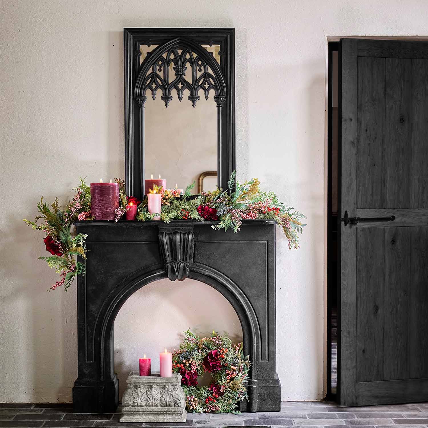 Cheminée noire en pierre avec miroir gothique, bougies bordeaux et roses entourées de décorations végétales vertes