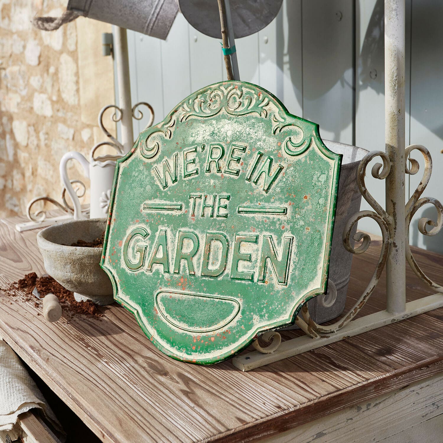 Panneau décoratif Merrin Plaque décorative en métal vert avec inscription We're in the Garden posée sur une table en bois dans un jardin