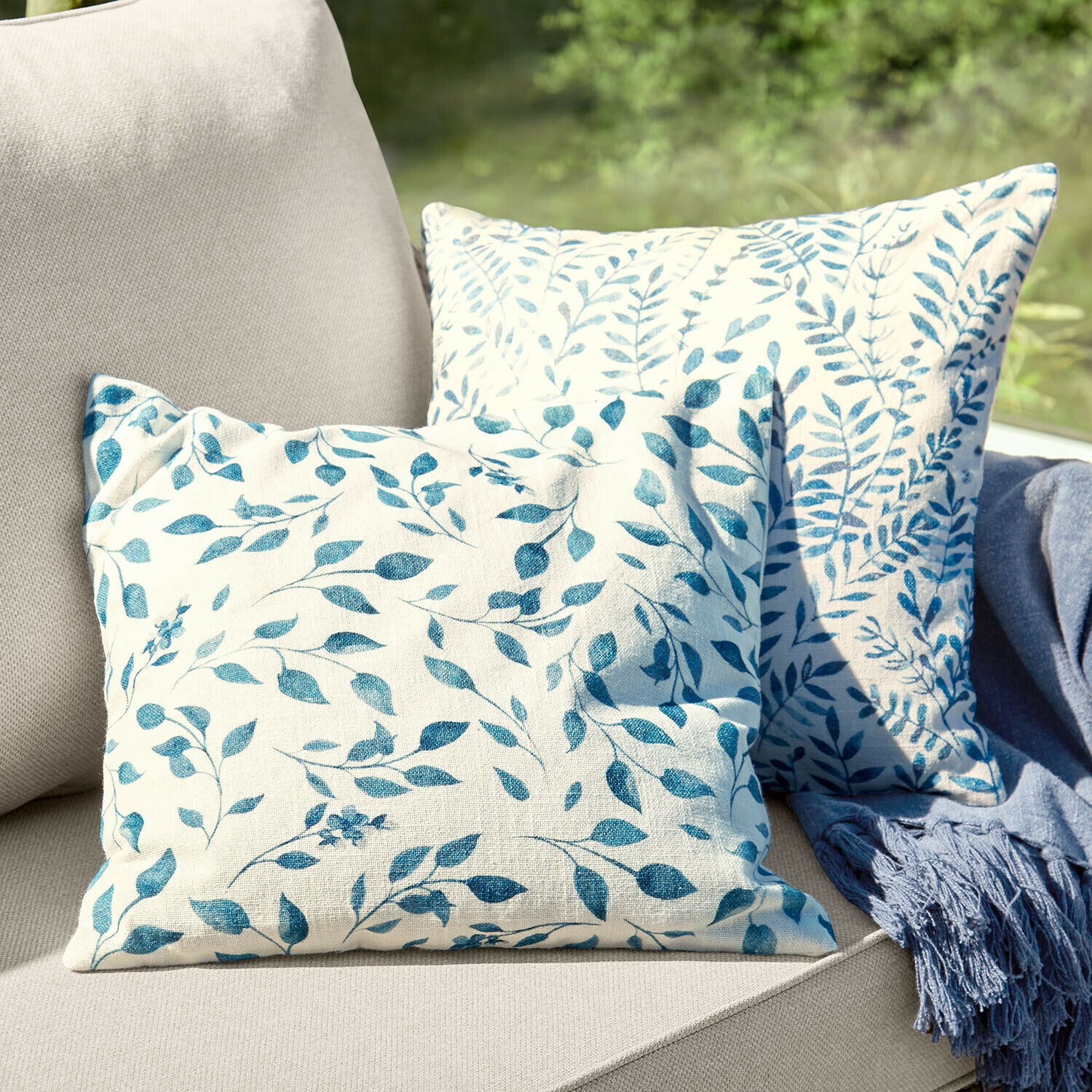 Lot de 2 housses de coussins Ocevara Deux coussins décoratifs en coton crème avec motif de feuilles bleues sur un canapé beige