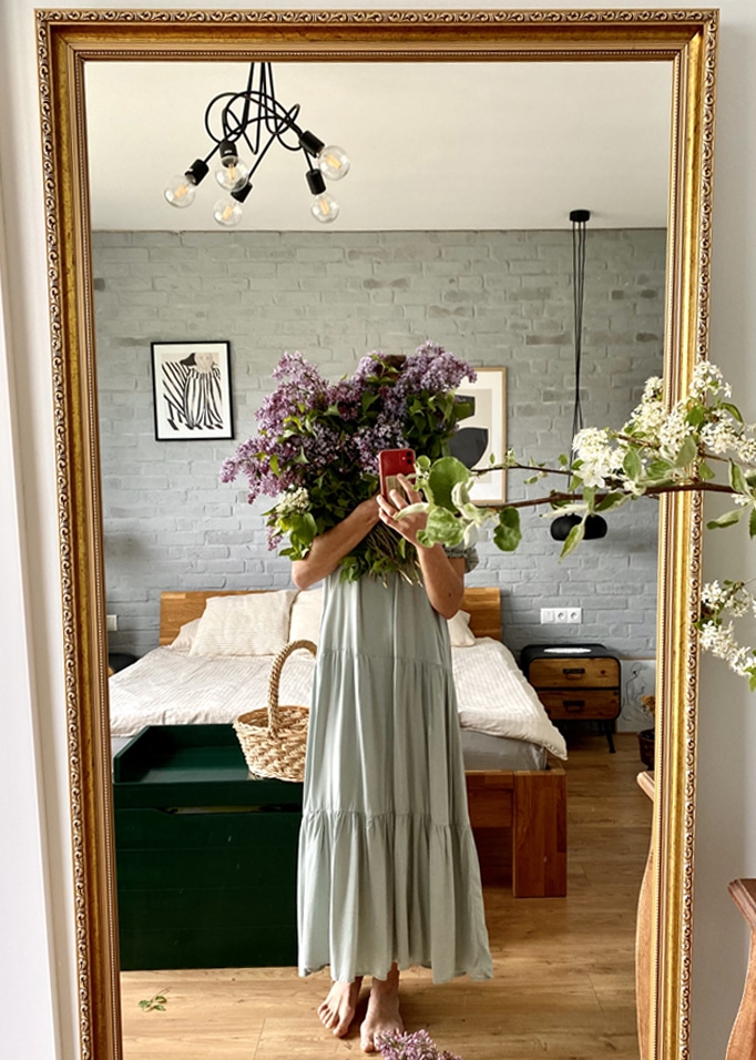 Femme en robe longue verte tenant un bouquet de fleurs violettes devant un miroir dans une chambre avec lit en bois
