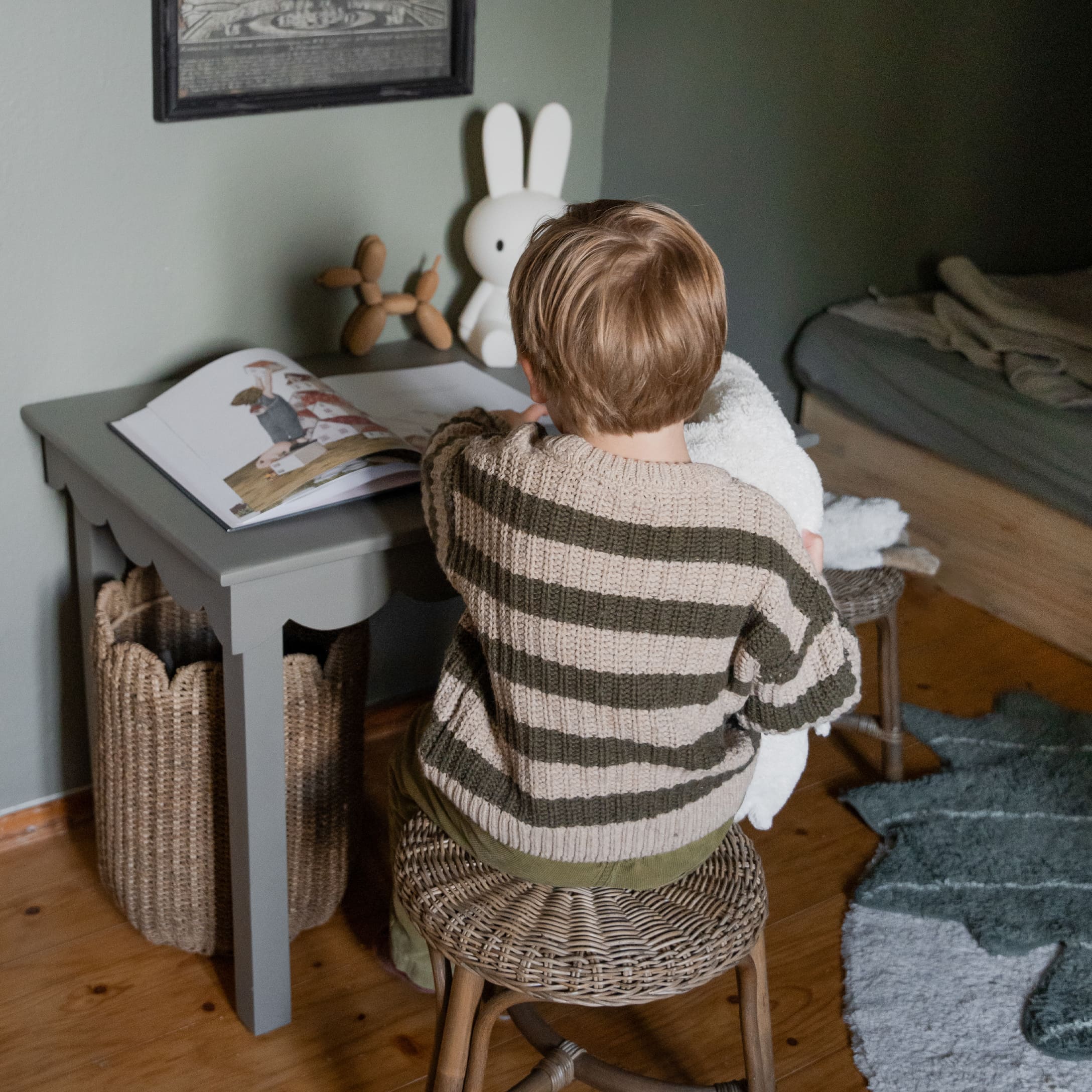 Enfant en pull rayé assis sur un tabouret en osier devant un bureau gris avec livre et décorations
