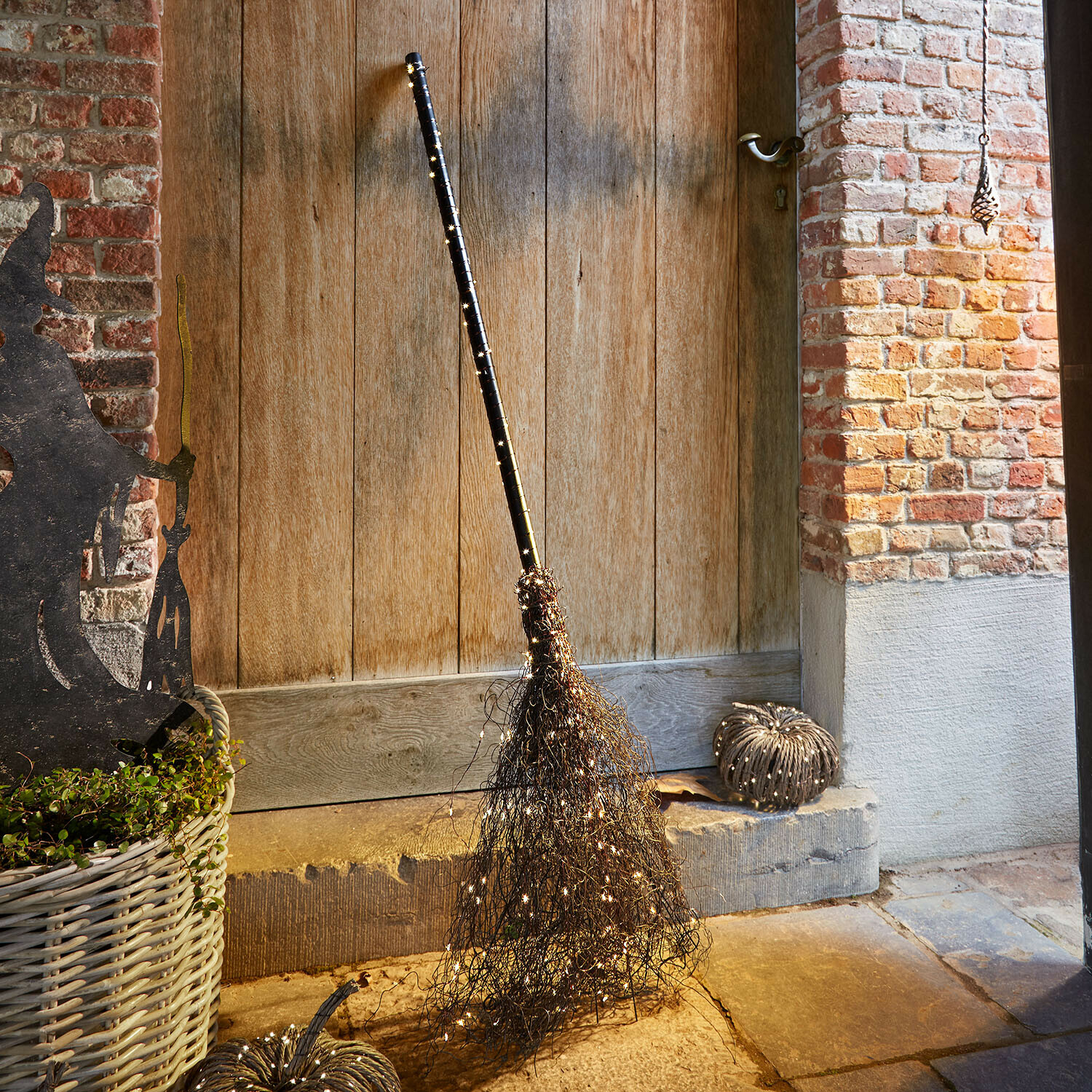 Balai décoratif Talmora Balai décoratif avec manche noir et guirlande lumineuse LED devant une porte en bois et un mur en briques