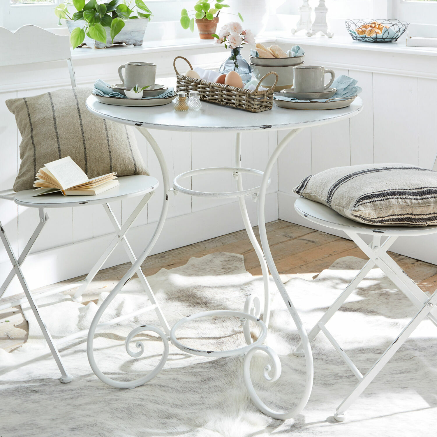 Table Iteuil Table ronde en métal blanc avec deux chaises pliantes et coussins décoratifs dans un intérieur lumineux