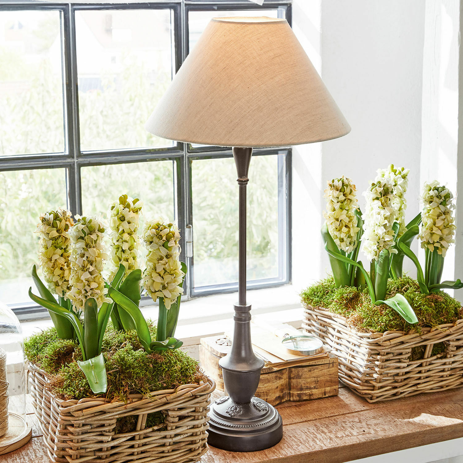 Lampe de table en métal avec abat-jour beige à côté de paniers en osier avec fleurs blanches et mousse