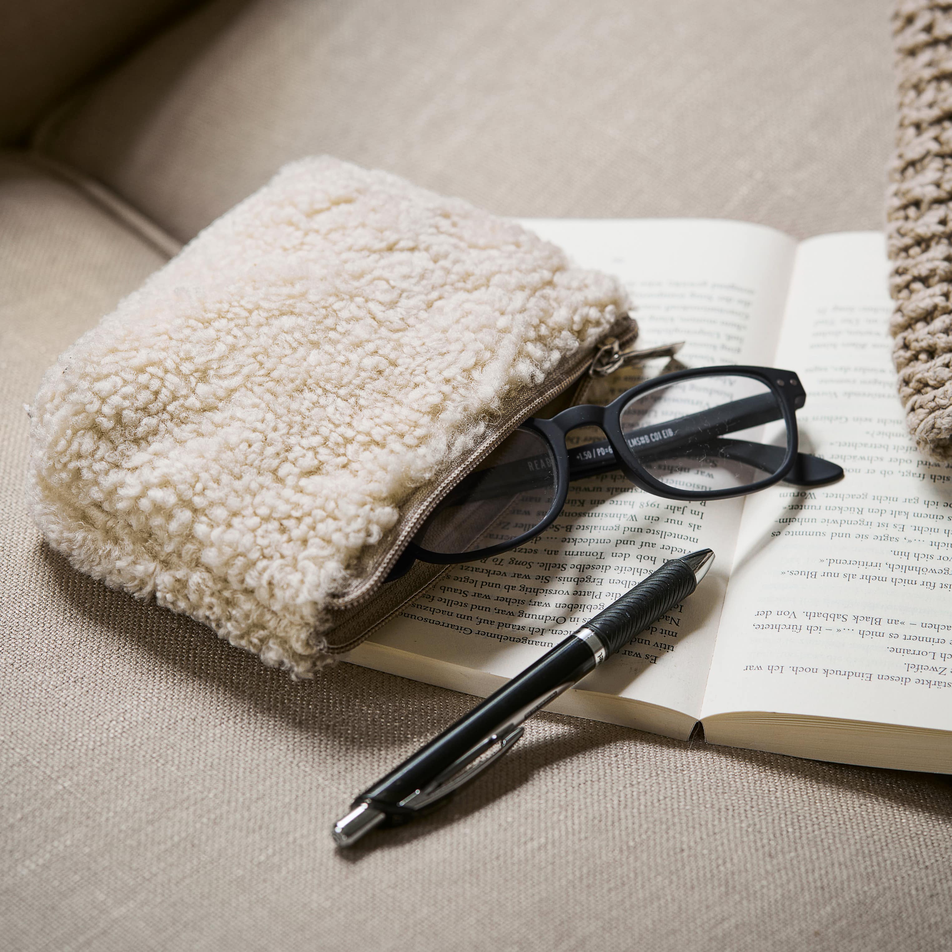Trousse beige en tissu pelucheux posée sur un canapé avec lunettes et stylo sur un livre ouvert