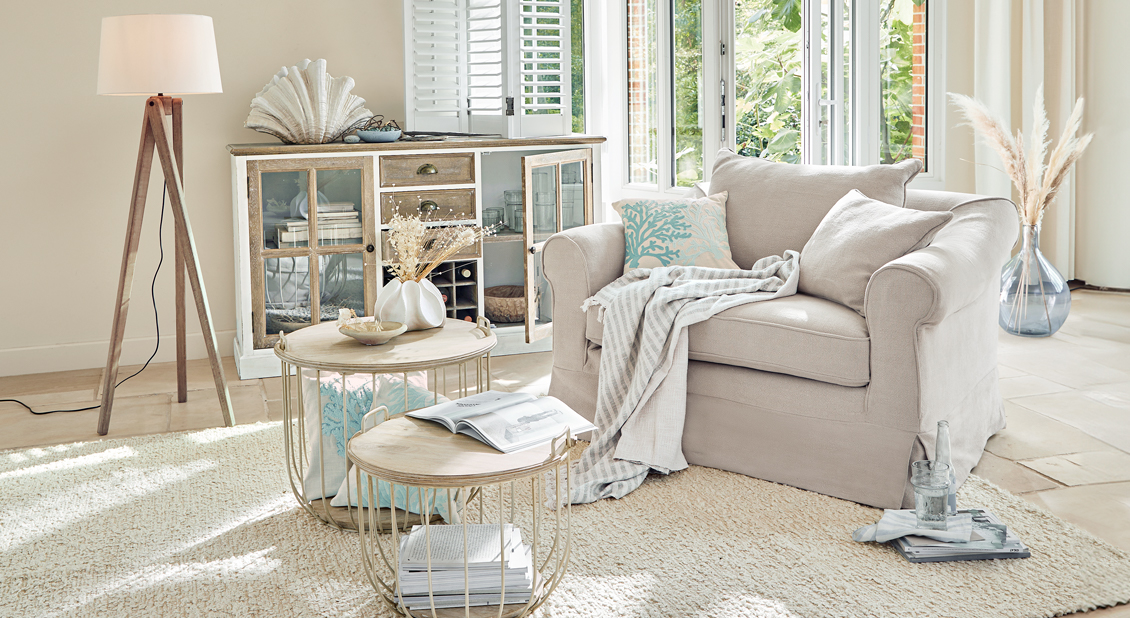 Fauteuil beige confortable avec coussins et plaid dans un salon lumineux avec tables en bois et lampe sur pied