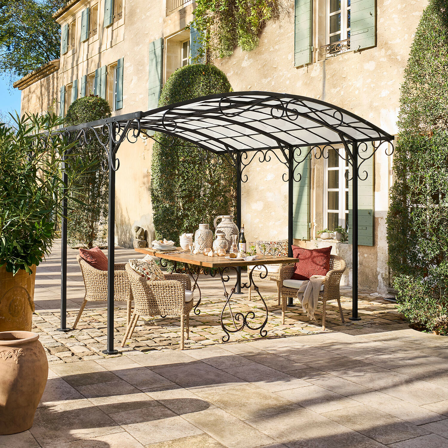 Pergola en métal noir avec décorations en volutes au-dessus d'une table en bois et chaises en osier