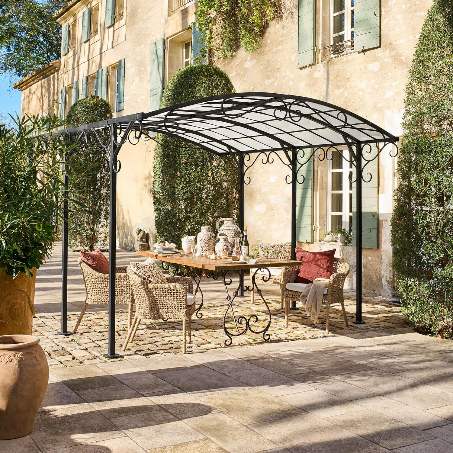 Pergola Delbridge Pergola en métal noir avec décorations en volutes au-dessus d'une table en bois et chaises en osier