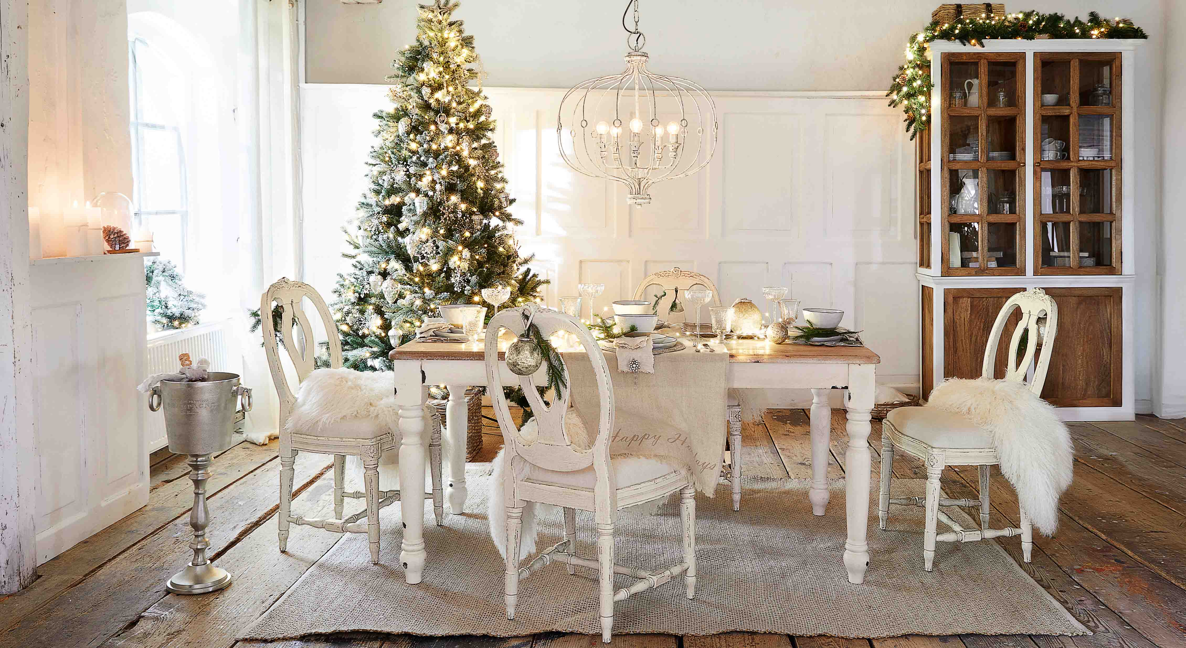 Salle à manger avec table et chaises en bois blanc décorée pour Noël avec sapin et guirlandes lumineuses