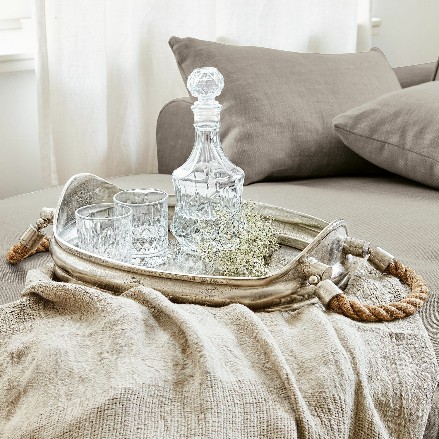 Plateau Morgan Plateau argenté avec poignées en corde, carafe en cristal et deux verres sur une couverture beige