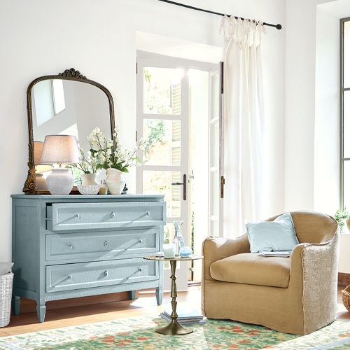 Commode en bois bleu clair avec miroir et décorations dans un salon lumineux avec fauteuil beige