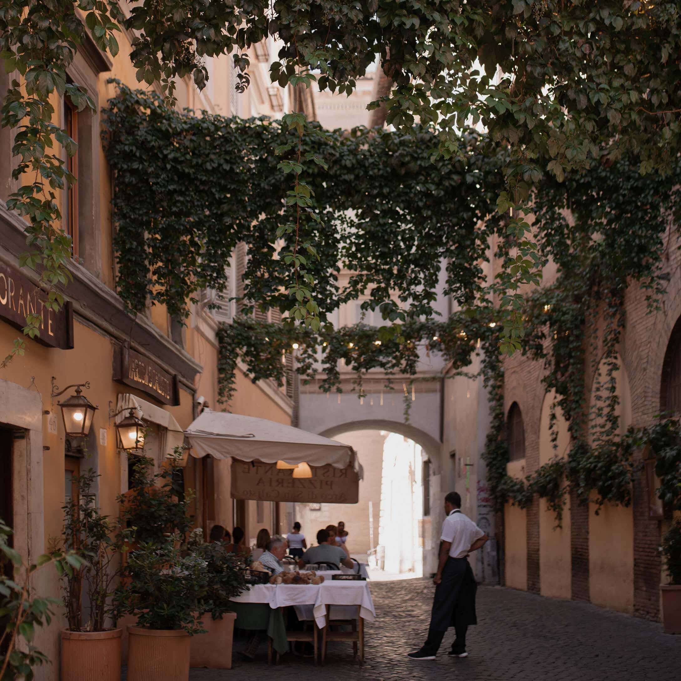Ruelle pavée avec restaurant en terrasse sous une pergola de verdure, ambiance chaleureuse
