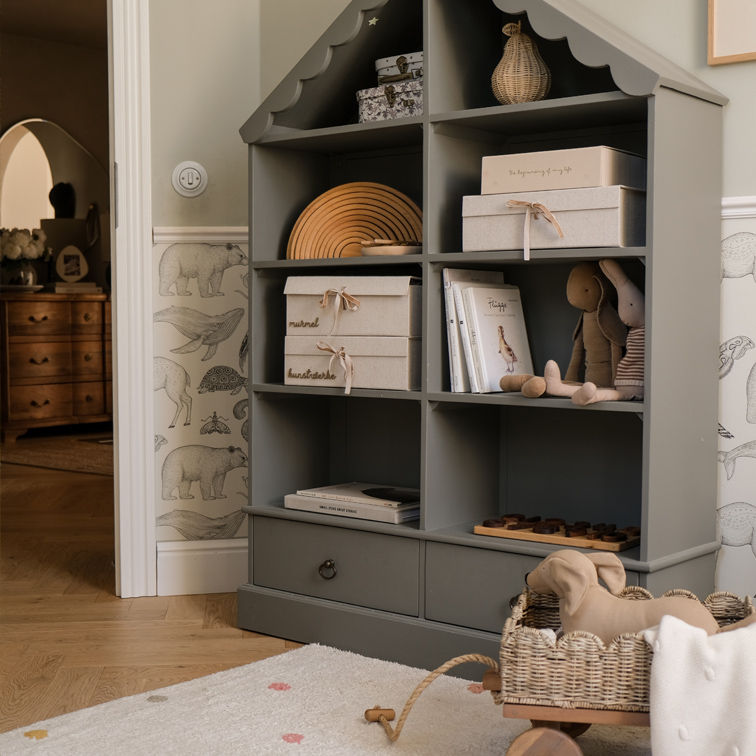 Étagère grise en bois en forme de maison avec boîtes, livres et peluches dans une chambre d'enfant