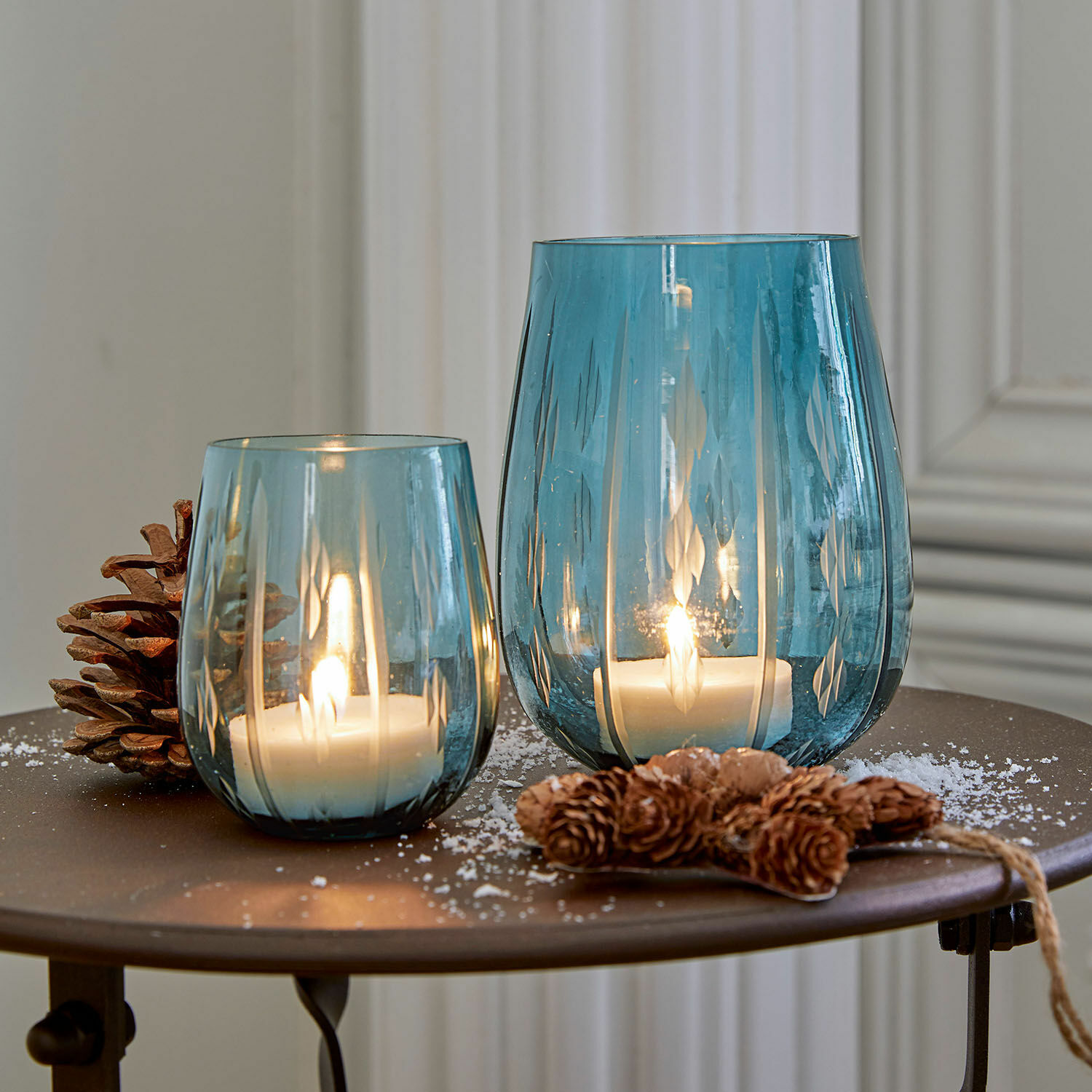 Lot de 2 photophores Danisar Deux photophores en verre bleu avec motifs, bougies blanches allumées et décoration de pommes de pin