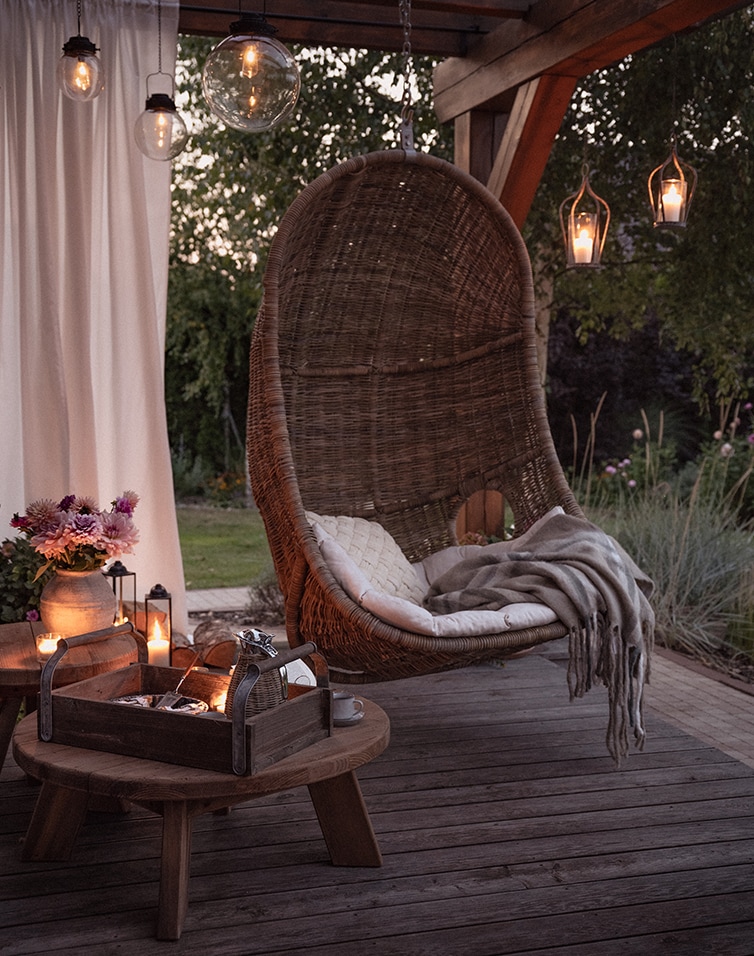 Fauteuil suspendu en rotin marron avec coussin et plaid sur terrasse en bois éclairée par des lampes décoratives