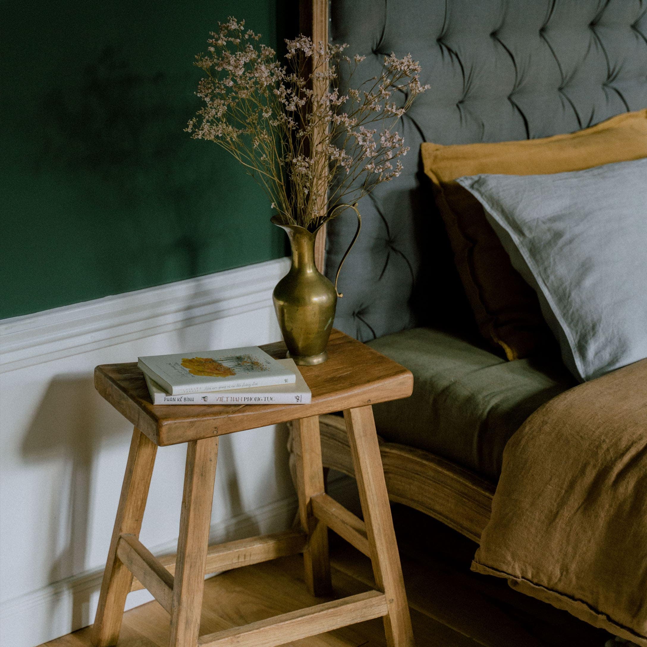 Tabouret en bois avec un pichet en laiton et des fleurs séchées à côté d'un lit avec tête de lit grise et literie marron