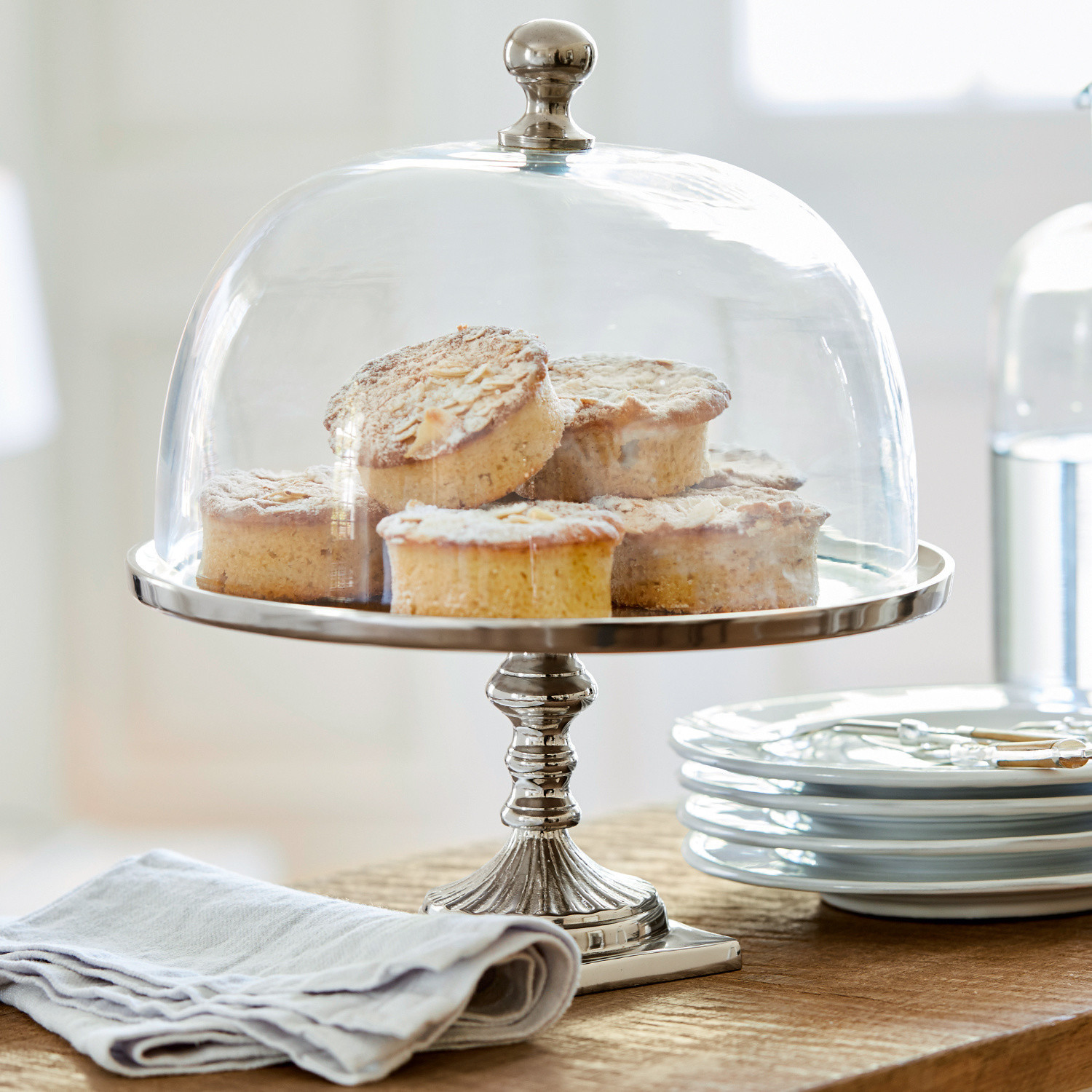 Cloche en verre avec plateau Mine Présentoir en métal avec cloche en verre transparent contenant des gâteaux sur une table en bois avec assiettes blanches