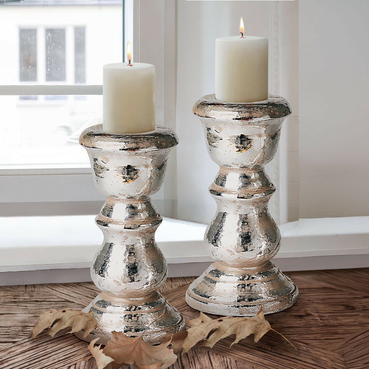 Lot de 2 bougeoirs Brilomine Deux bougeoirs argentés avec bougies blanches sur un sol en bois près d'une fenêtre, décoration automnale