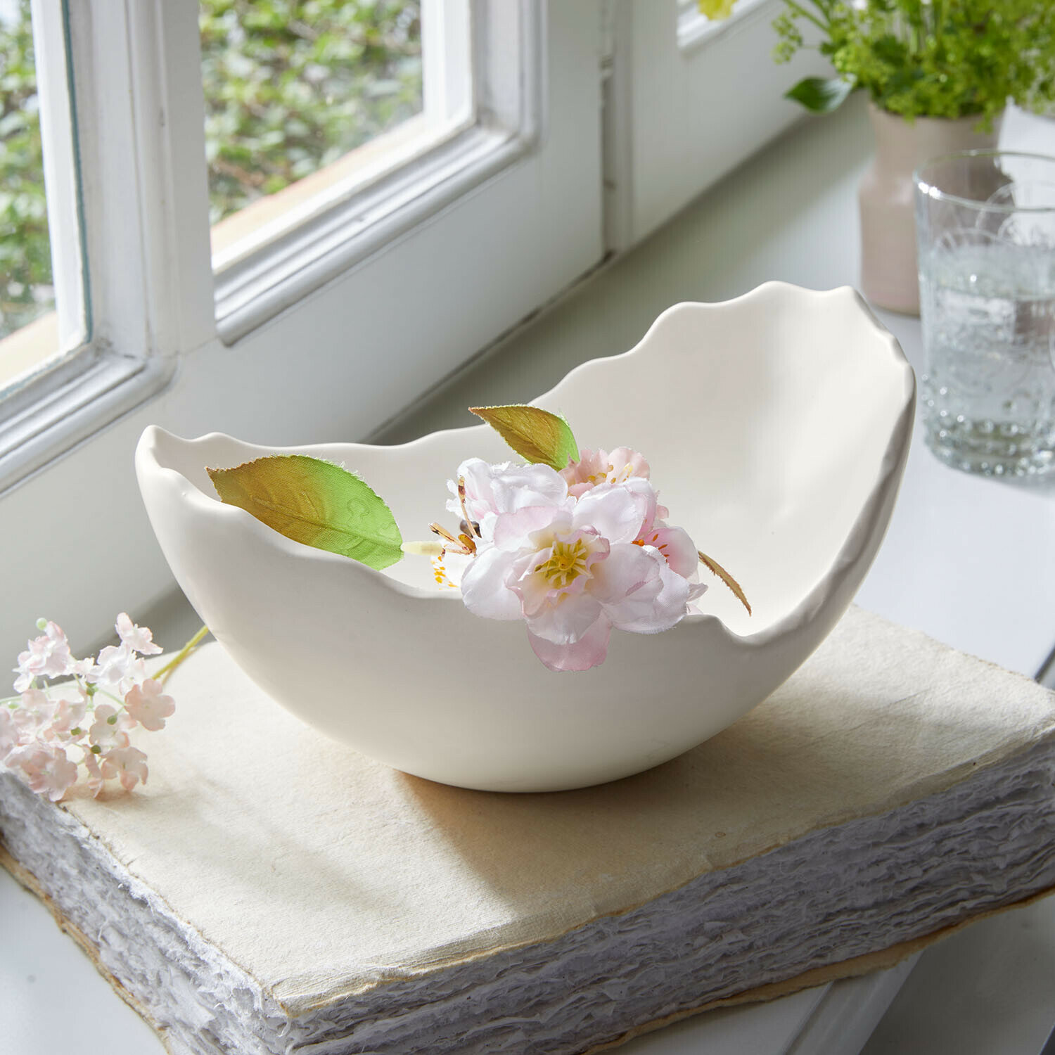 Coupe Aureen Bol en céramique blanche à bords ondulés avec une fleur délicate posé sur un livre près d'une fenêtre