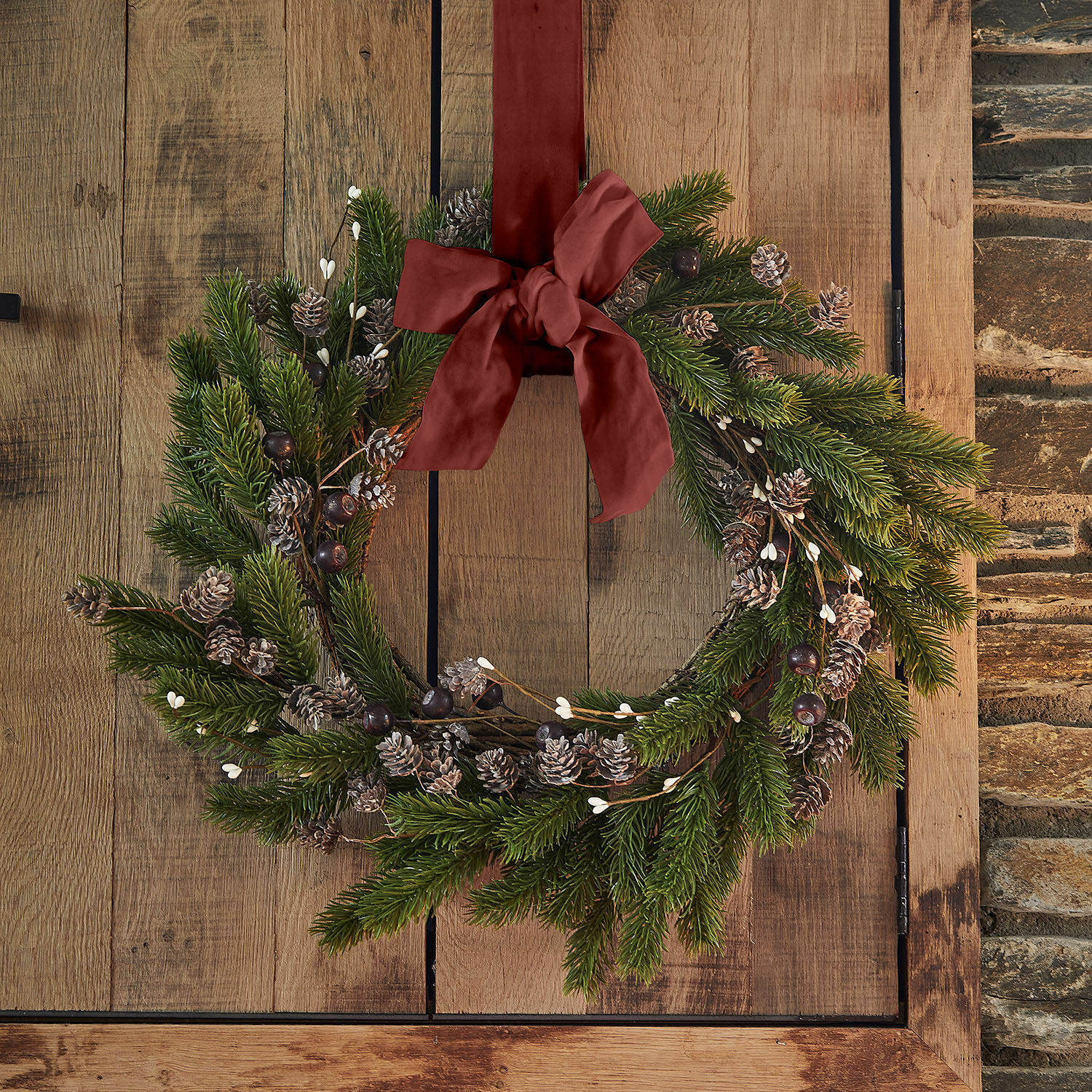 Couronne Lyandra Couronne de Noël en branches vertes et pommes de pin avec un nœud bordeaux sur une porte en bois